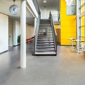 Light grey concrete effect safety flooring in a large corridor with a metal staircase and yellow panel walls